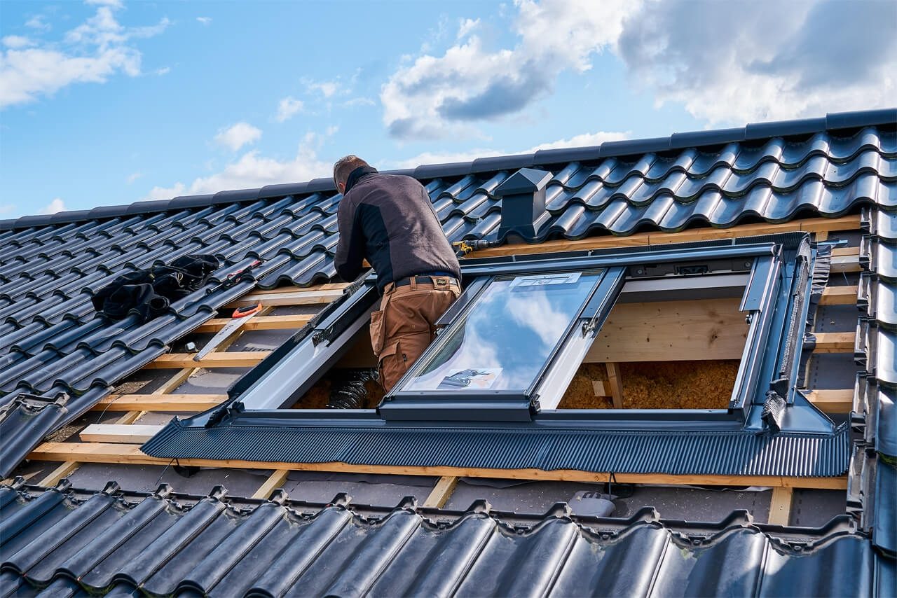 A fitter on a tiled roof installing a VELUX roof window: tiles stripped back to the
          battens, the window seated in its opening and the grey flashing kit being dressed in
          around it.