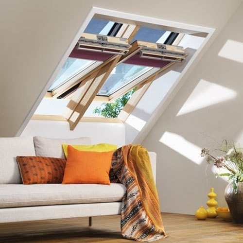 Two pine-finished VELUX centre-pivot roof windows set into the sloping ceiling of a
          loft room, both propped open, with sunlight falling across a sofa below.